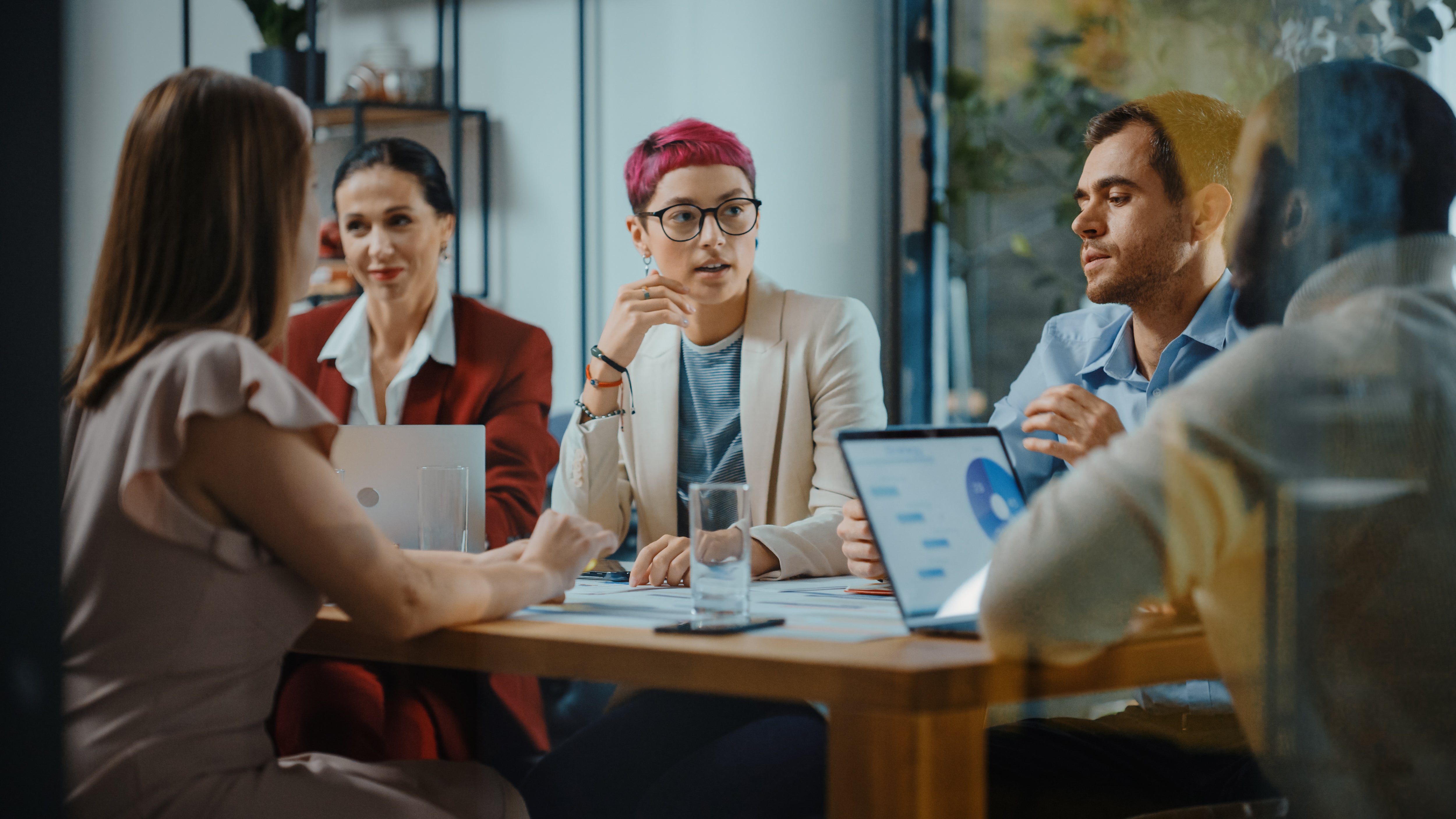 Five people in a meeting room, discussing while looking at laptops and documents. A woman with pink hair speaks. Office setting, focused atmosphere.