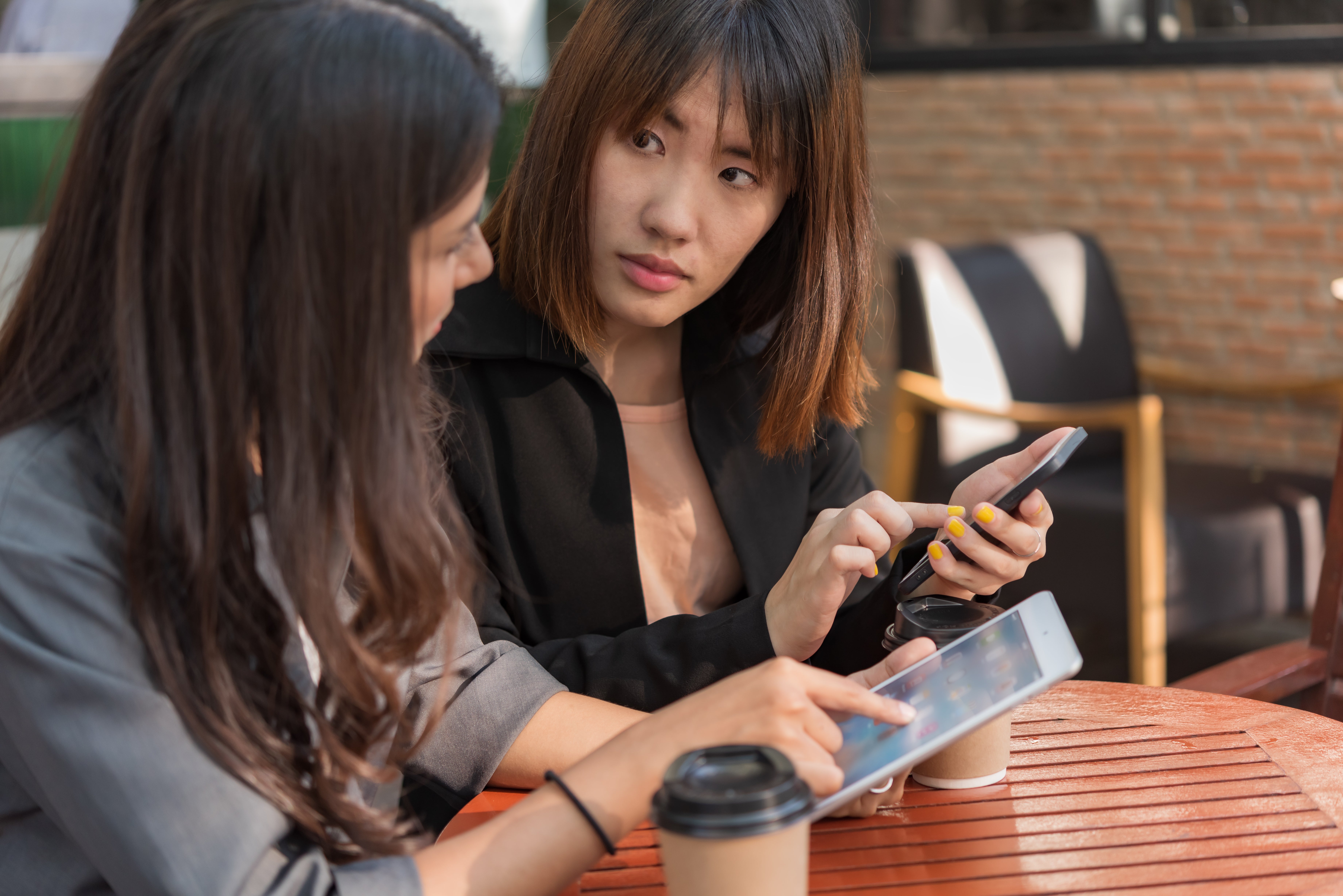 Two women at a cafe table using a phone and tablet. One looks focused, with yellow nails. Coffee cups on table. Background is blurred.