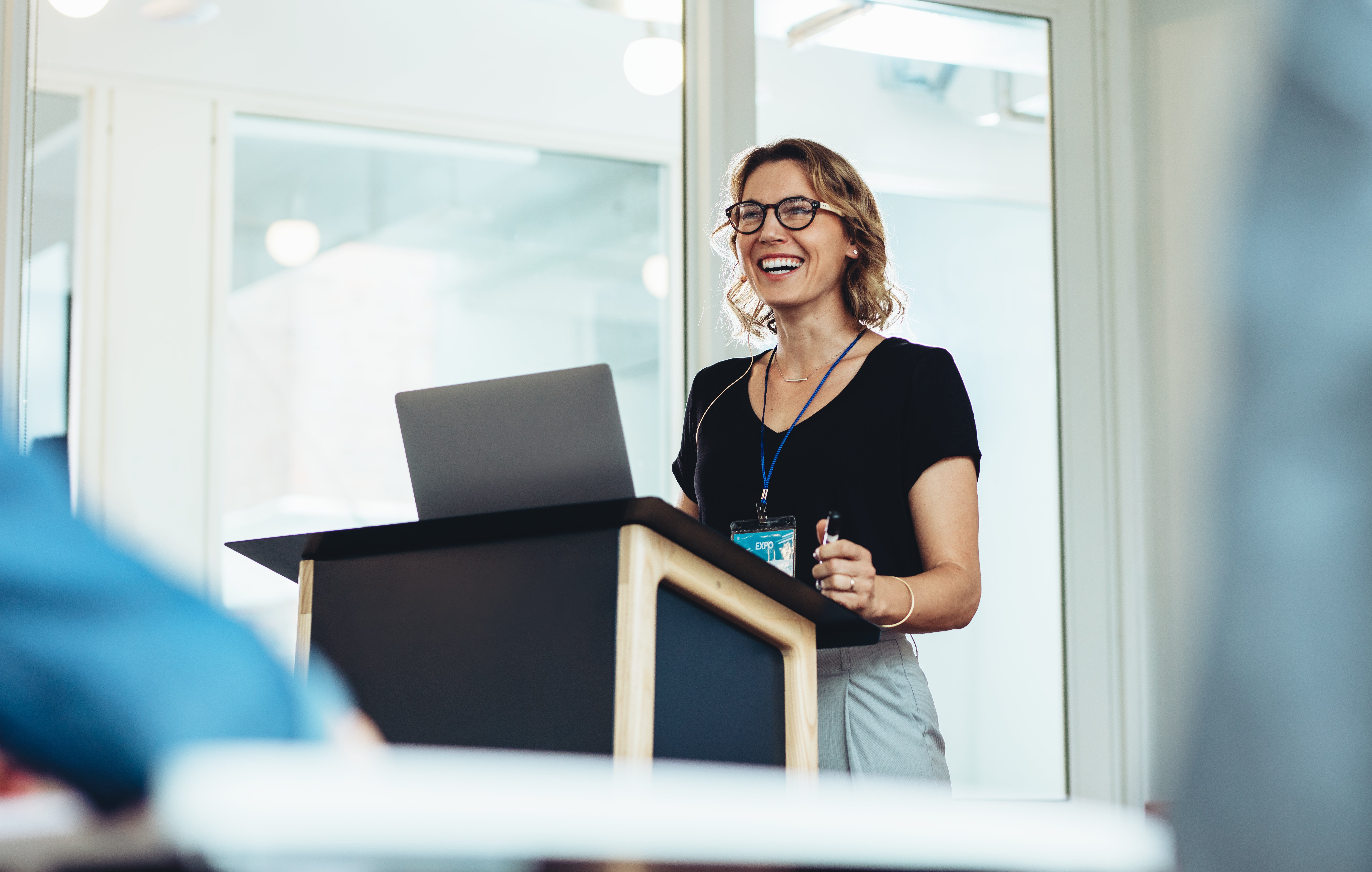 Woman smiling at a podium with laptop, holding a microphone. She's wearing glasses and a badge. Glass-walled room, bright atmosphere.