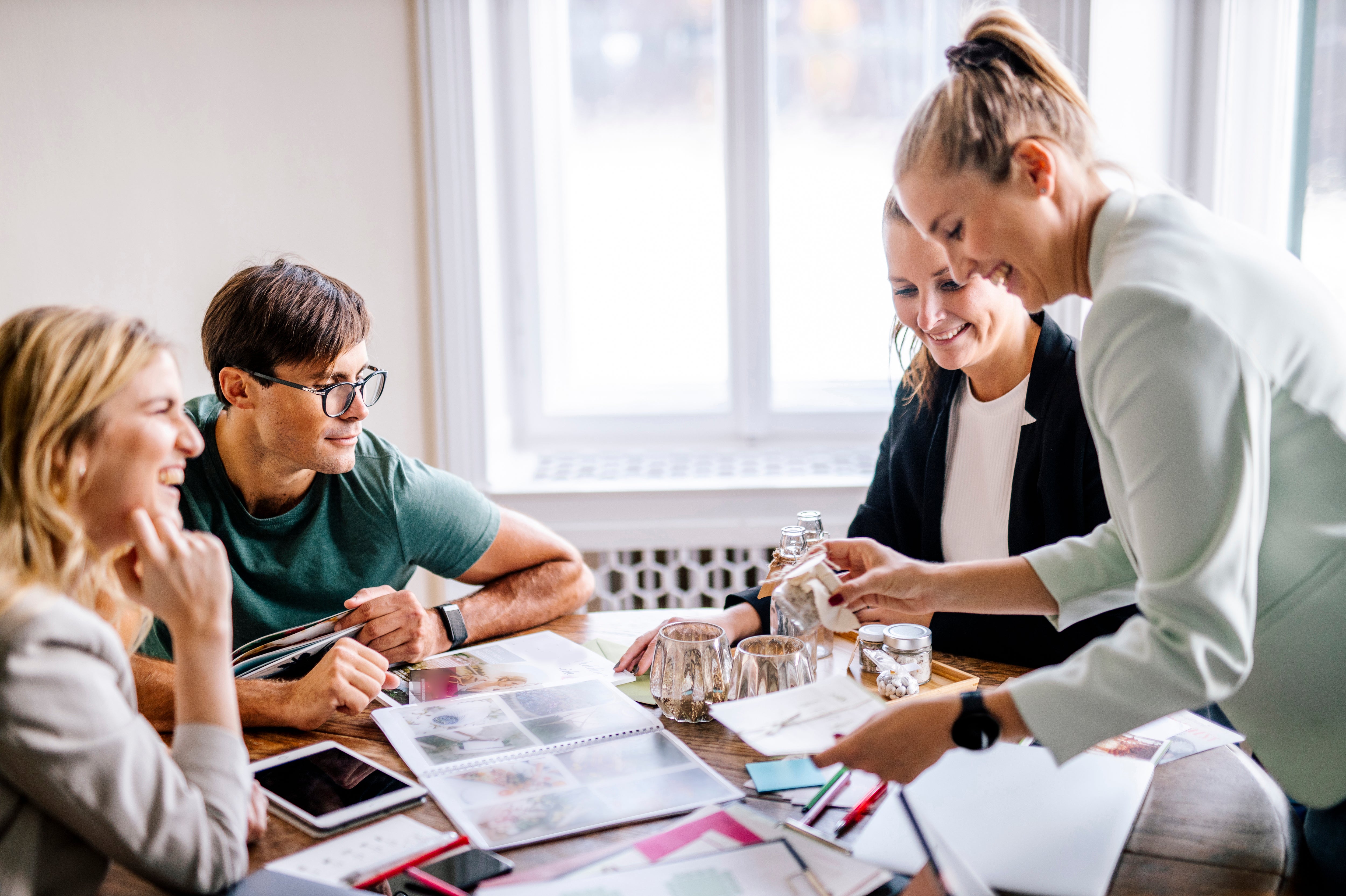 Four people collaborate happily around a table with colorful papers and notebooks, bright indoor setting with a large window.