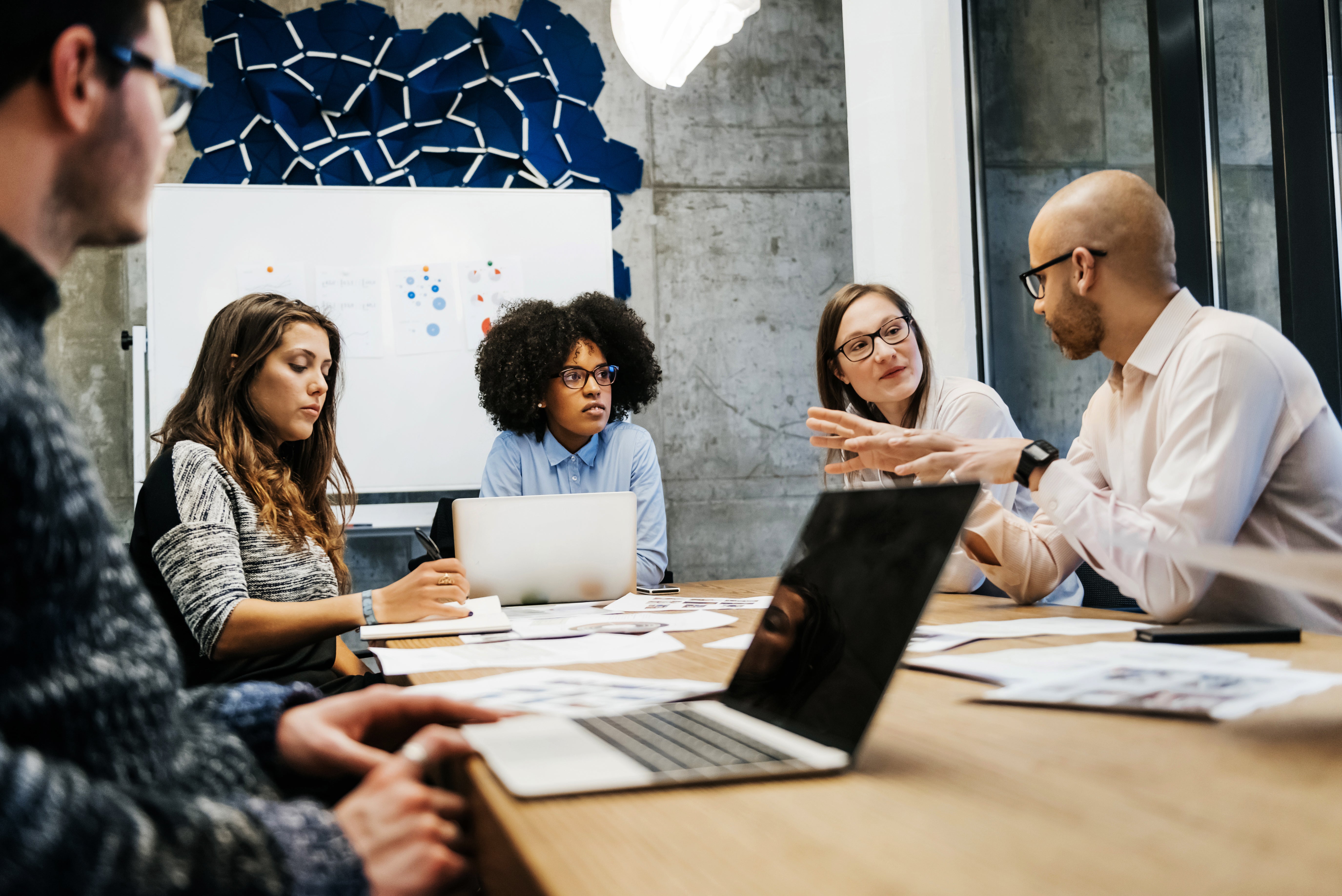 Five people in a meeting discussing ideas around a table with laptops. Abstract blue art on a concrete wall background. Engaged and focused mood.