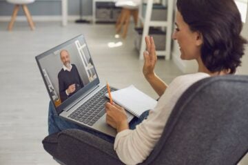 : A woman sits in a chair with a laptop on her legs, while a man on her screen discusses business English lessons