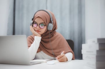A young woman sits at her laptop with headphones engaged in a business English training course.