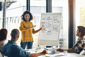 A business professional in a yellow sweater presenting a plan in front of colleagues