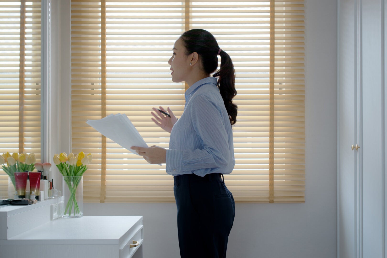 A woman in a blue dress shirt practicing speaking in front of a mirror holding papers and a pen