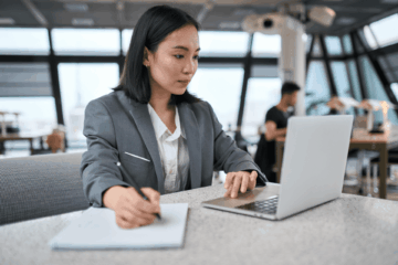 An office worker working on a laptop while taking notes on a piece of paper