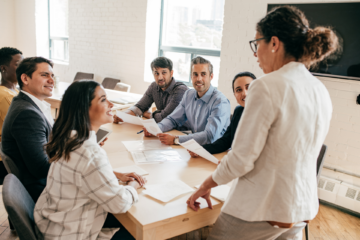 A manager stands at the head of a wooden table leading a meeting with her team