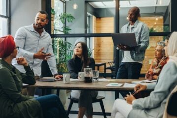 Team meeting in modern office, five diverse people engaged in discussion. One man gestures while another holds a laptop. Casual atmosphere.