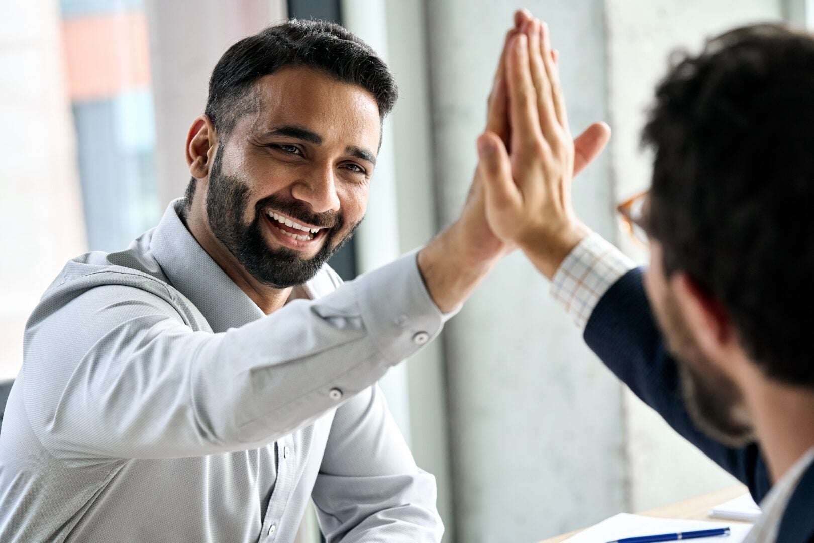 Two men in an office high-fiving, smiling. One wears a light shirt, the other a dark suit. Bright, modern background. Happy mood.