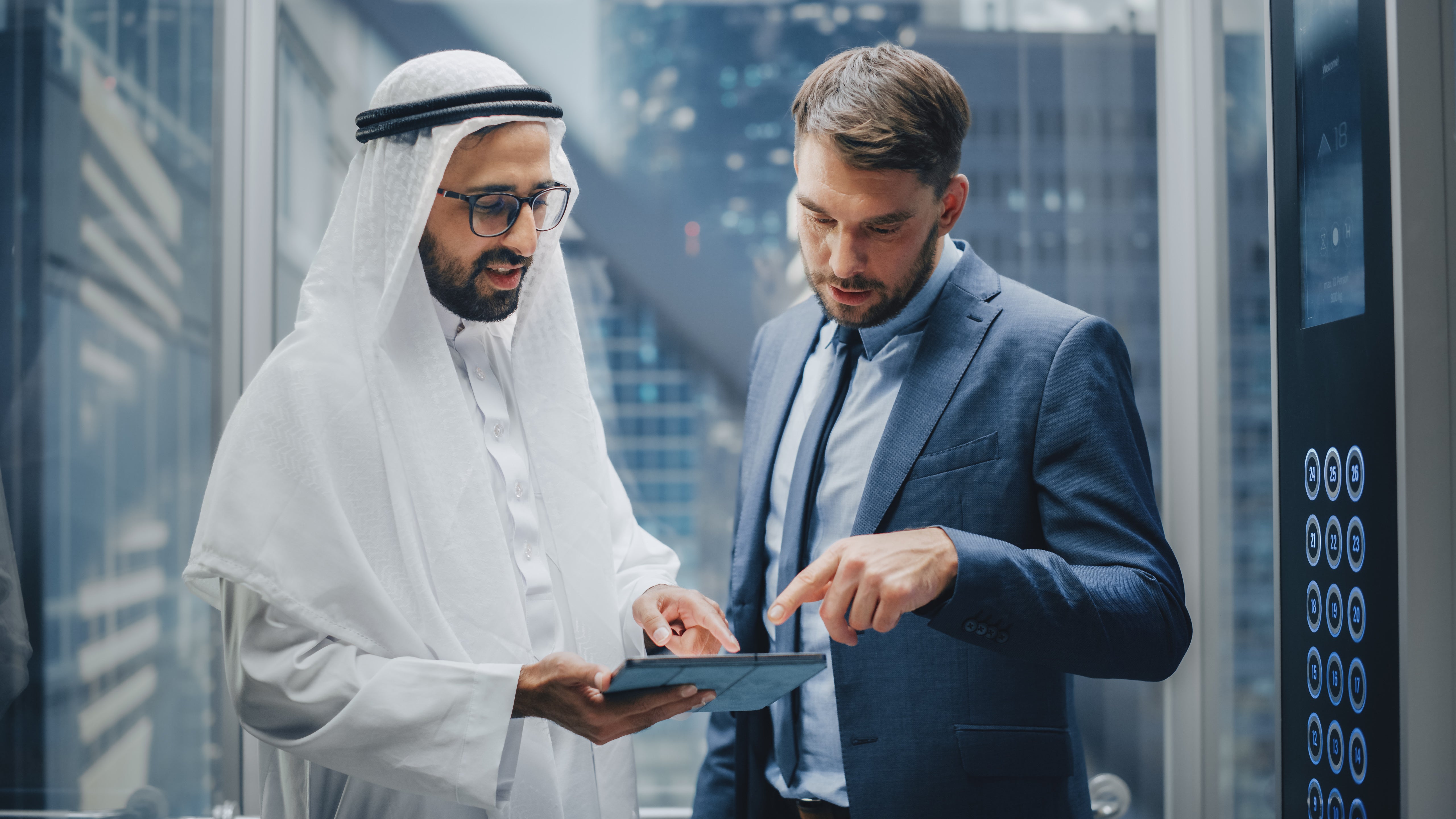 Two men in formal attire discuss something on a tablet in an elevator. One wears a kandura; the other a suit. Cityscape visible outside.