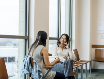 Two women sit and talk in a bright room with large windows. One holds notes, while the other listens, creating a thoughtful atmosphere.