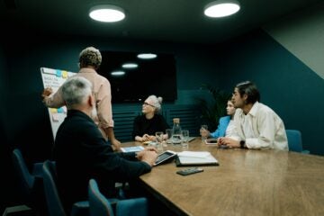 Man presents data to four colleagues in a bright office. They review charts on paper and laptop, engaged in discussion. Plant visible.