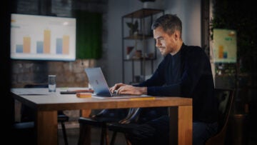 Man working on laptop at wooden table in dimly lit room. Background: blurred charts on screen, shelves, greenery. Focused and calm mood.