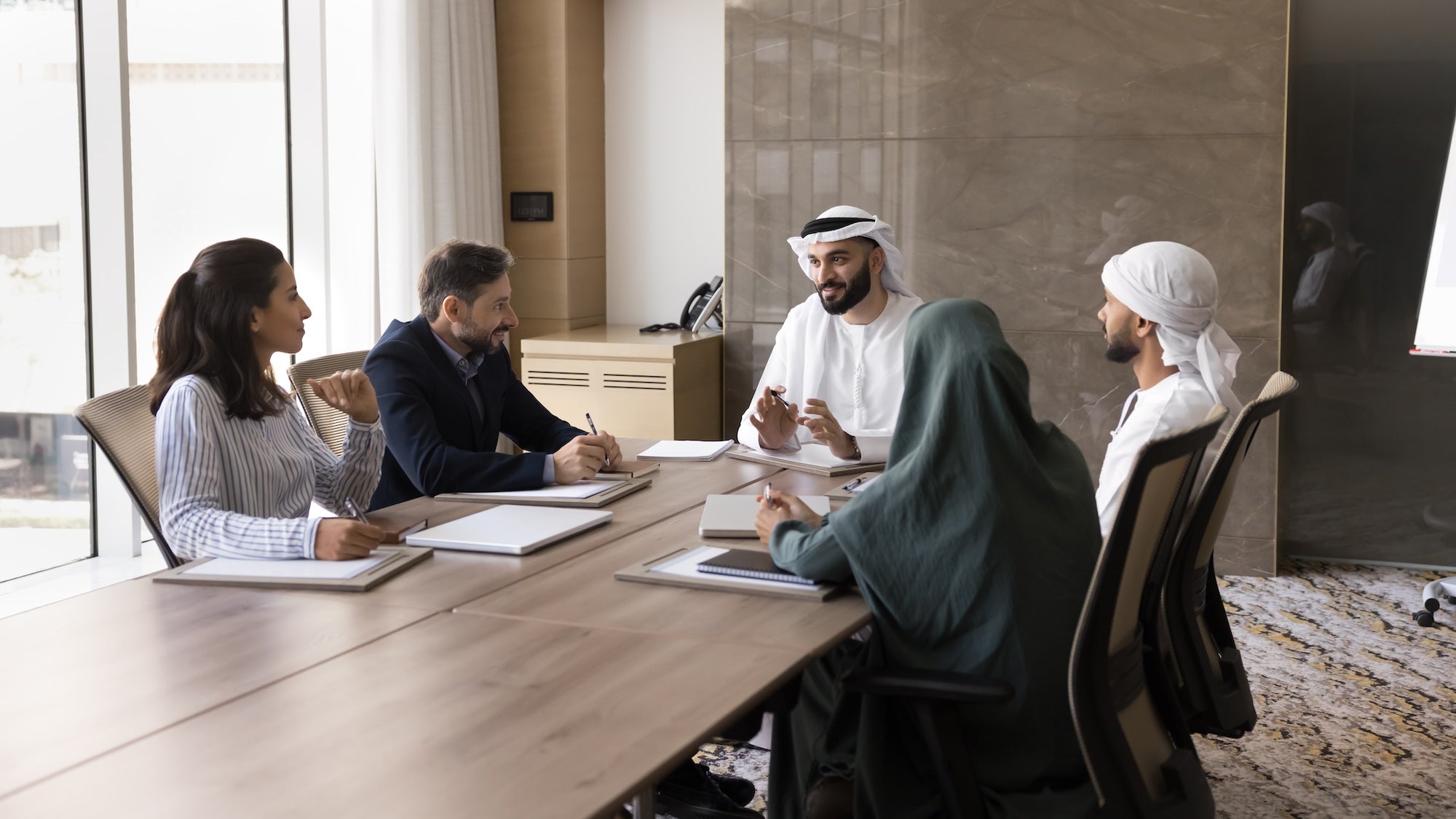 Five people in a meeting, discussing around a table in a bright office. Notebooks and pens on table, with a collaborative atmosphere.