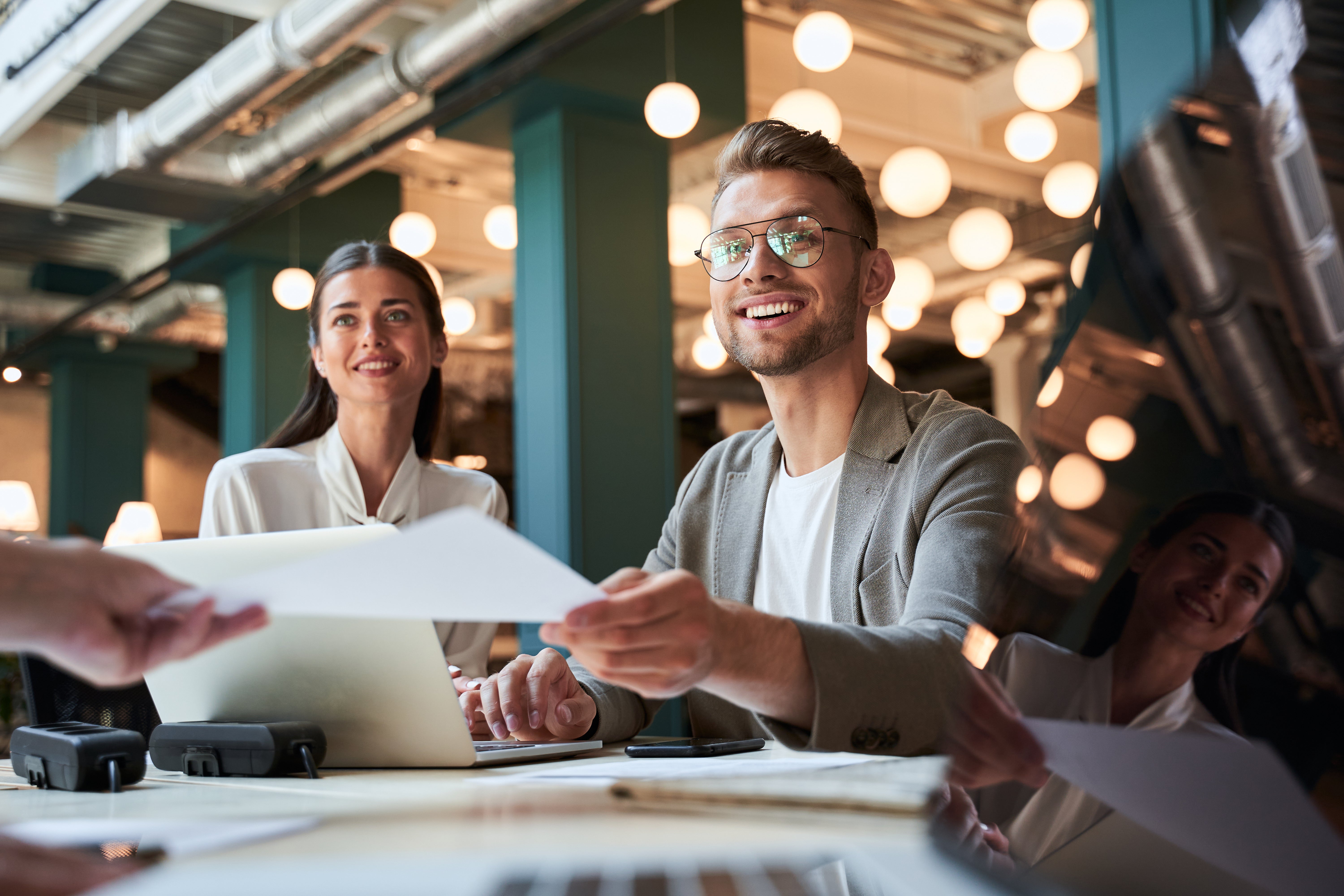 Two smiling people in a modern office setting, exchanging documents. Warm lighting and industrial decor create a collaborative atmosphere.