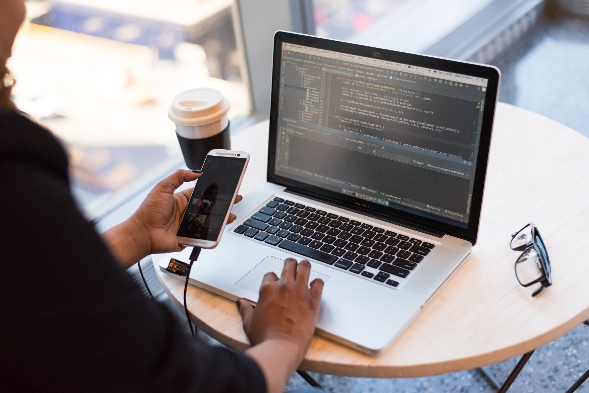 Person at a round table using a laptop displaying code, holding a smartphone, with glasses and a coffee cup nearby.