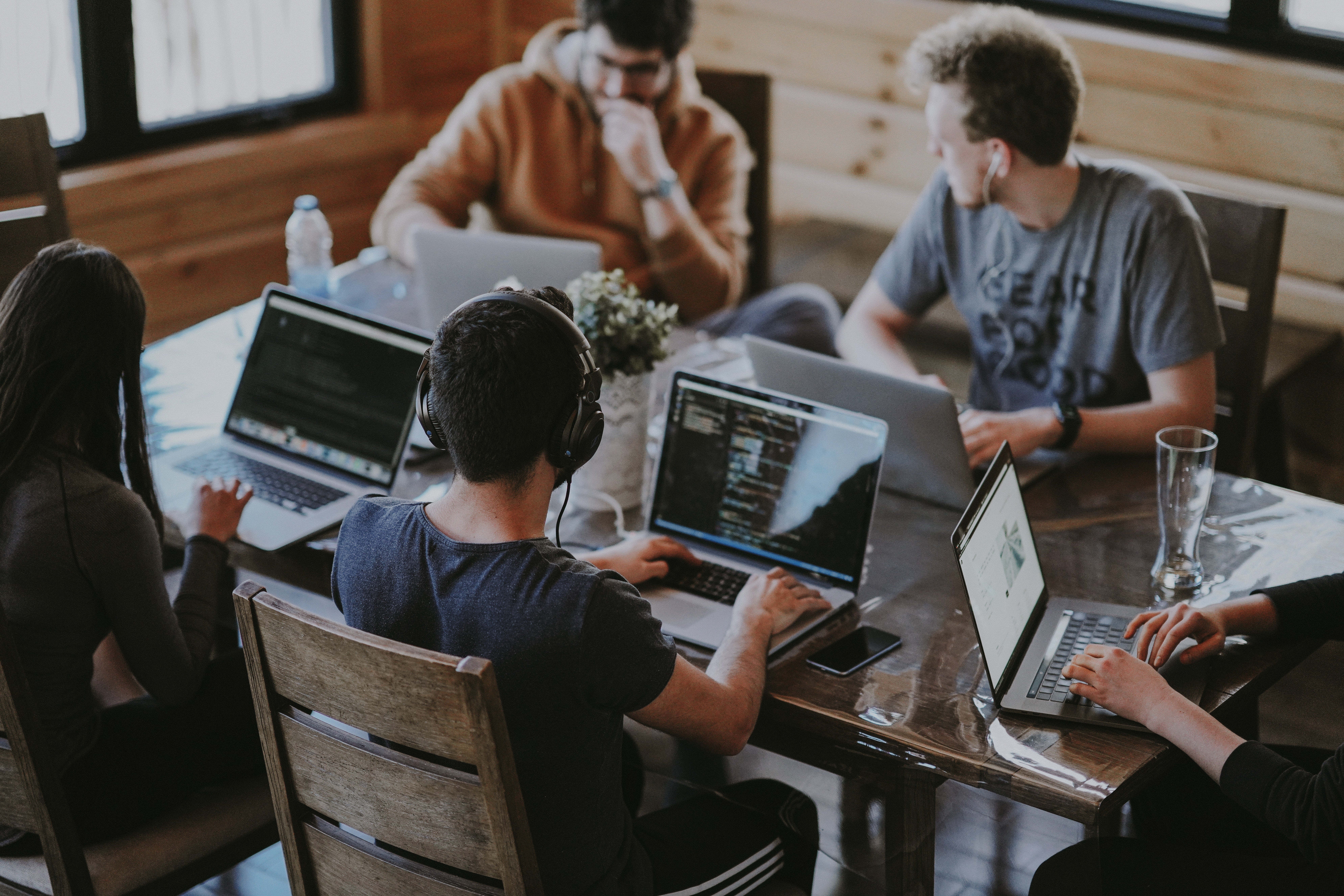 Five people working on laptops around a wooden table, wearing casual clothes, focused atmosphere, natural light from windows, plants on table.