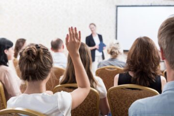 Audience in a seminar room, one woman raising hand. Presenter in focus at front by a screen. Mood is attentive and engaged.