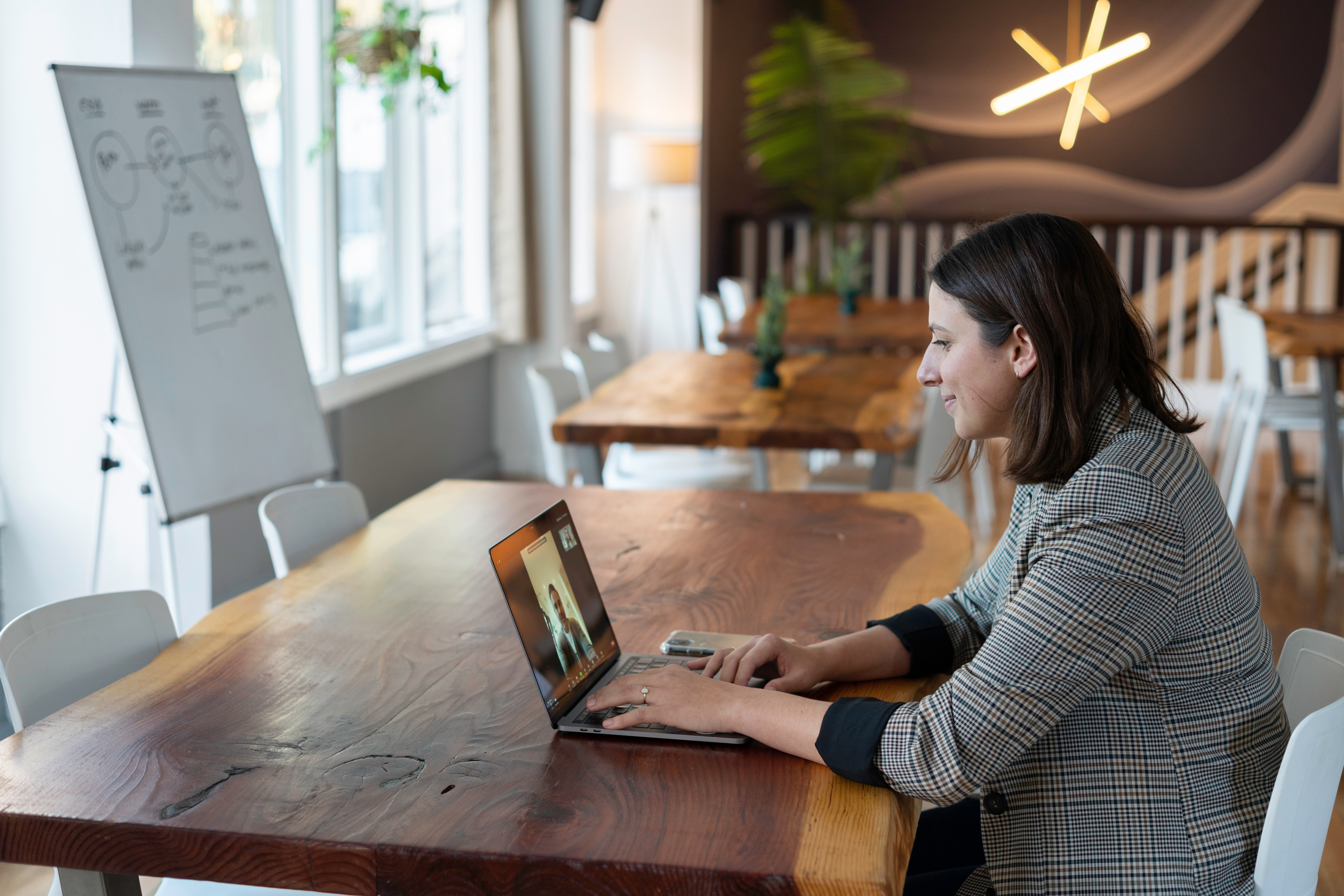 Woman on a video call using a laptop in a bright room with wooden tables and a whiteboard. Casual, focused atmosphere.