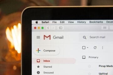 Close-up of a laptop screen displaying Gmail in Safari browser. Inbox is highlighted in red. Warm light bulb glows in the blurred background.