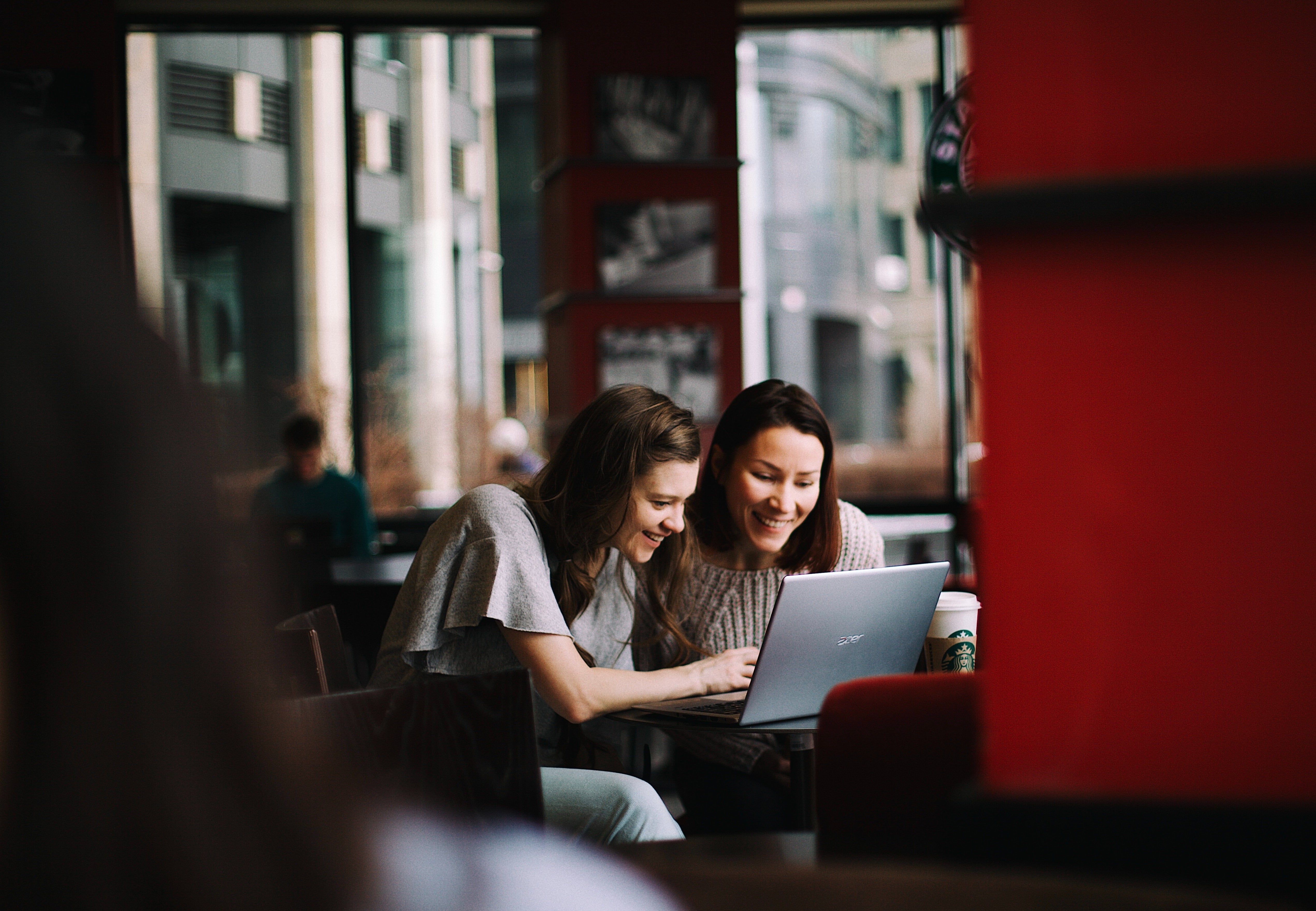 Two women happily look at a laptop in a café with large windows. A Starbucks cup is on the table. The atmosphere is cozy and modern.