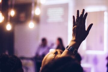 Hand raised in focus with people blurred in the background, ambient glowing lights. The setting appears to be a dimly lit event or meeting.