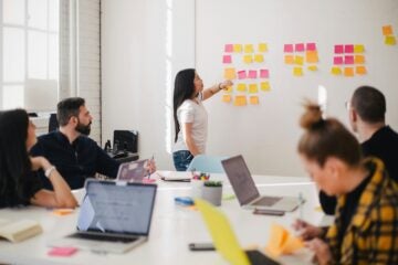 Woman in white shirt presenting sticky notes on a whiteboard to a group. Laptops and notes on table. Bright room, collaborative mood.