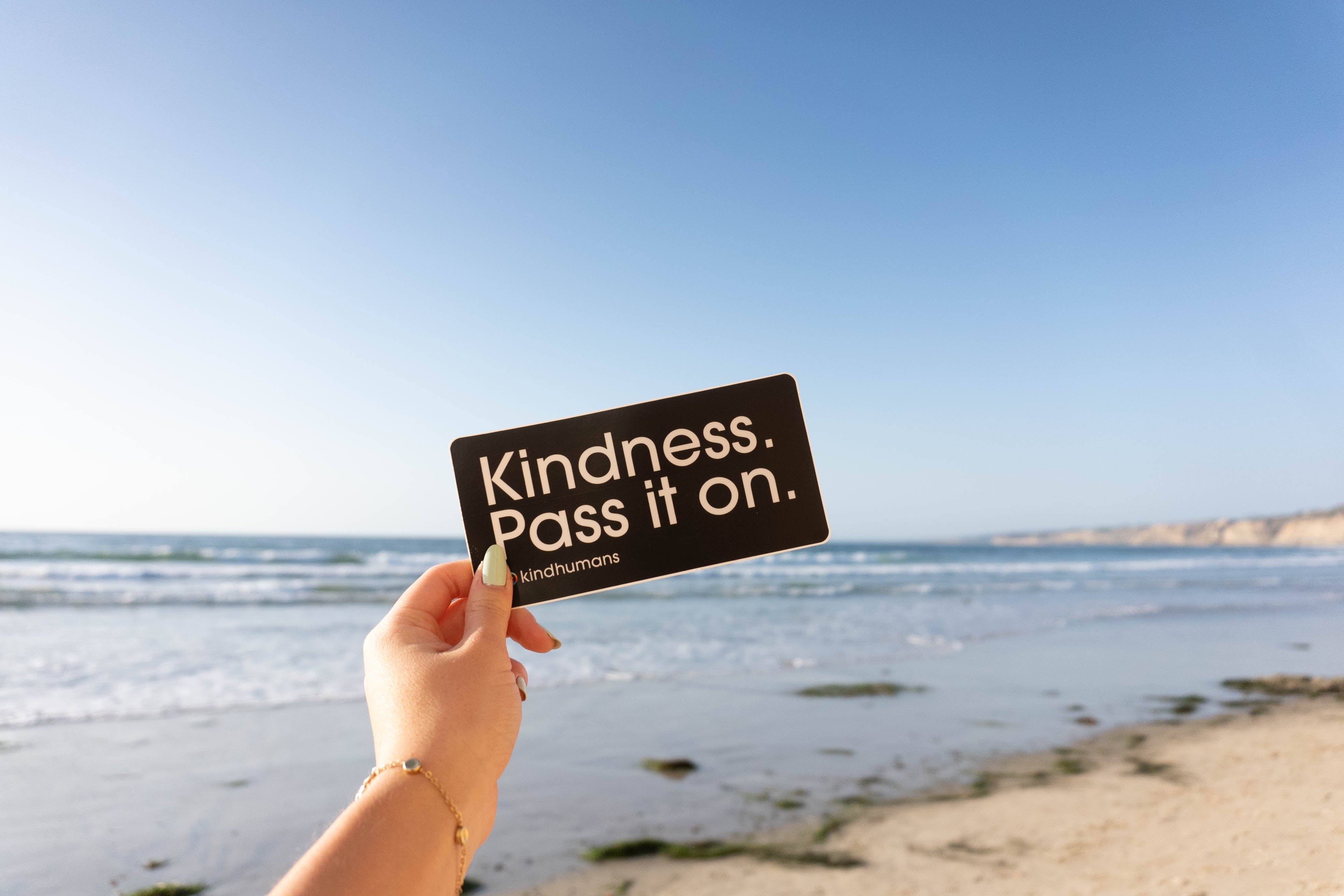 Hand holding a black card saying "Kindness. Pass it on." over a sunny beach background with waves and clear blue sky.